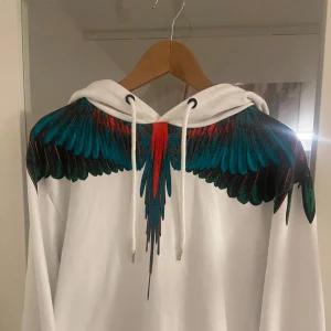  Marcelo Burlon hoodie - Säljer en vit hoodie från Marcelo Burlon med ett  vingmönster i blått, rött och svart över axlarna. Hoodien är i nyskick och har använts fåtal gånger ✅ skriv för frågor eller byten⚡️💬