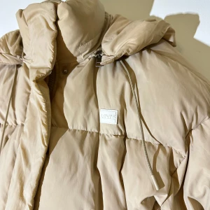 Beige pufferjacka från Levi's - Säljer en snygg beige pufferjacka från Levi's. Jackan har en hög krage och dragsko vid huvan för extra skydd mot kyla. Perfekt för kalla dagar med sin vadderade design. Levi's-logga på bröstet ger en stilren touch.