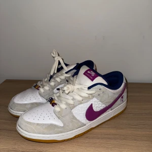 Nike SB Dunk Low - Säljer ett par Nike SB Dunk Low med vit och lila färgkombination. Skorna har en vit bas med lila detaljer och en gummisula. De är utrustade med snörning och har en klassisk Nike-logga på sidan.