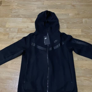 Svart hoodie från Nike - Säljer en stilren svart hoodie från Nike med dragkedja och huva. Jackan har långa ärmar och diskreta detaljer i form av svarta band över bröstet och ärmarna. Perfekt för en sportig look eller avslappnad stil. Den kommer också med byxor