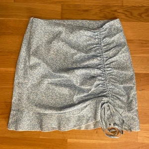 Blommig blå kjol med snörning - Söt blå kortkjol med ett blommigt mönster och snörning på sidan. Kjolen har en volangdetalj längst ner som ger en extra touch. På sidan finns en dragkedja för att enkelt få på sig den. Perfekt för en sommaren!💞