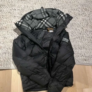 Svart dunjacka från Burberry - Säljer en snygg svart dunjacka från Burberry med avtagbara ärmar. Jackan har en klassisk rutig insida och en huva. Taggs finns.
