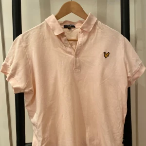 Ljusrosa pikétröja från Lyle & Scott - Säljer en ljusrosa pikétröja från Lyle & Scott med klassisk krage och knappslå. Tröjan har korta ärmar och ett broderat emblem på bröstet. 