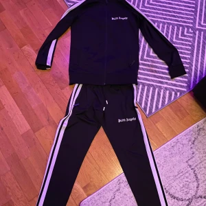 Svart tracksuit från Palm Angels - Snyggt svart träningsset från Palm Angels med vita ränder längs ärmar och ben. Jackan har dragkedja och byxorna har elastisk midja. Perfekt för en sportig look.