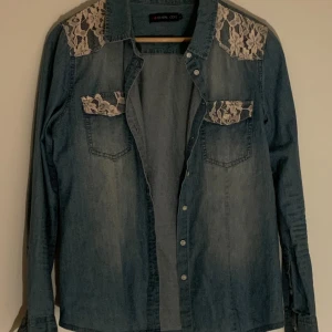 Jeansskjorta med spetsdetaljer från Even&Odd - Snygg jeansskjorta från Even&Odd med spetsdetaljer på axlar och fickor. Skjortan har långa ärmar och knappar framtill. Perfekt för en trendig look med en touch av elegans. Pris går att diskutera vid snabb affär. Kontakta gärna mig vid frågor och funderingar.