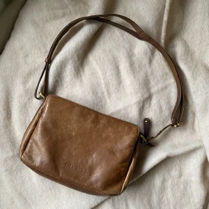 Brun väska i läder - Jättefin mindre väska i äkta läder från märket The Monte. Bandet är justerbart så den funkar både som axelväska, handväska och axelremsväska (crossbody). I jättefint skick och har många fack både med dragkedja och utan, och hela väskan stängs med en dragkedja.