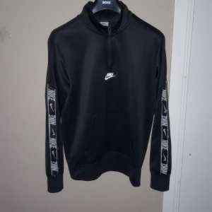 Svart tröja från Nike - Säljer en svart tröja från Nike med dragkedja vid halsen och vita detaljer. Ärmarna har ett mönster med Nike-loggor i vitt och svart. Tröjan är storlek s 