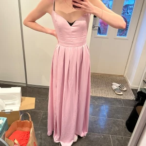 Ljusrosa balklänning - Elegant ljusrosa balklänning med smala axelband och veckad kjol. Klänningen har en stilren design med en öppen rygg och korsade band. Perfekt för speciella tillfällen.