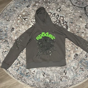 Sp5der Hoodie - Säljer en grå hoodie från Sp5der med ett coolt spindelnätmotiv och gröna detaljer på framsidan. Tröjan har långa ärmar och en bekväm huva. Perfekt för en avslappnad stil.