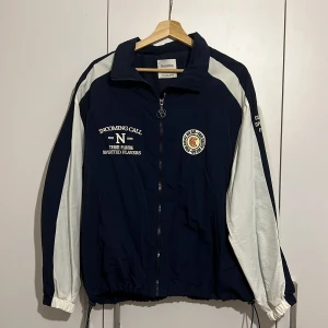 Blå och vit vindjacka från Bershka - Säljer en blå och vit vindjacka från Bershka med dragkedja och tryck på bröstet. Jackan har långa ärmar och en sportig look. Perfekt för en casual stil. 🌀använd en gång.