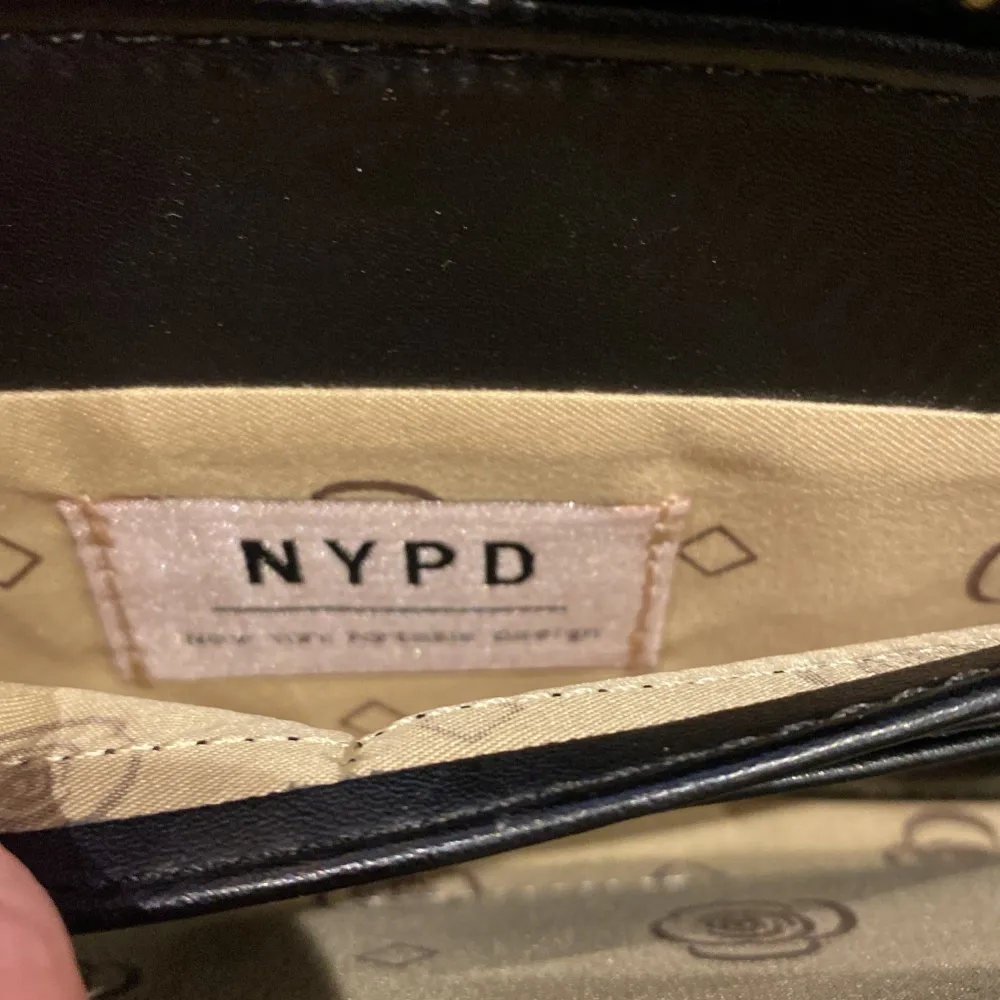 Snygg svartvit plånbok från NYPD med ett unikt mönster och dragkedja. Plånboken har flera fack för kort och en praktisk dragkedja på baksidan. Perfekt för att hålla ordning på dina tillhörigheter.. Laukut & Käsilaukut.