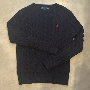 Mörkblå stickad tröja från Polo Ralph Lauren - Säljer en snygg mörkblå stickad tröja från Polo Ralph Lauren med det klassiska röda logotypbroderiet på bröstet. Tröjan har ett flätat mönster och långa ärmar, perfekt för kyligare dagar. Passar både till jeans och chinos!
