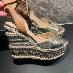 Glittriga kilklackar med remmar från River Island - Snygga kilklackar från River Island i glittrigt silver&guld med bruna remmar. Skorna har en hög platå med dekorativa detaljer och ett spänne runt ankeln. Perfekta för att ge en extra touch till din outfit. Inköpta i Miami