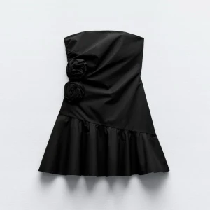 Svart axelbandslös klänning - Snygg svart axelbandslös klänning med volangdetalj och dekorativa blommor på sidan. Perfekt för en elegant kvällslook. Aldrig använd då den är helt ny, prislappen är kvar. Passade mig tyvärr inte