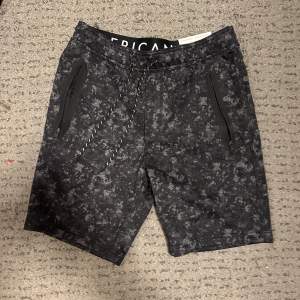 Snygga svarta joggershorts från American Eagle med ett diskret kamouflagemönster. De har praktiska dragkedjefickor och en bekväm snörning i midjan. Perfekta för en avslappnad stil.