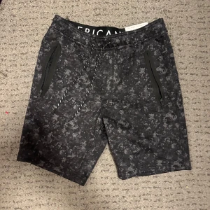 Svarta joggershorts med kamouflagemönster - Snygga svarta joggershorts från American Eagle med ett diskret kamouflagemönster. De har praktiska dragkedjefickor och en bekväm snörning i midjan. Perfekta för en avslappnad stil.