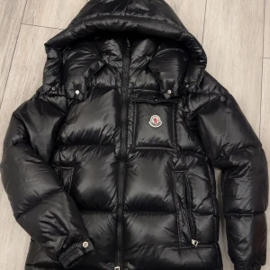 Svart pufferjacka från Moncler - Säljer en snygg svart pufferjacka från Moncler med huva och dragkedja. Jackan har ett stilrent Moncler-märke på bröstet och praktiska fickor. Perfekt för kyliga dagar när du vill hålla stilen.  Det finns ett litet hål vid vänster armlängds men det är inget som påverkar funktionen 🖤 bilden kan skickas om så önskans