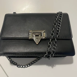 Svart axelväska med kedja - Snygg svart axelväska i skinn med en stilren kedja som axelrem som dessutom är justerbart. Väskan har ett metallspänne framtill som ger en cool touch. Perfekt för att bära dina nödvändigheter med stil. Köpt för 100kr säljs för 40kr