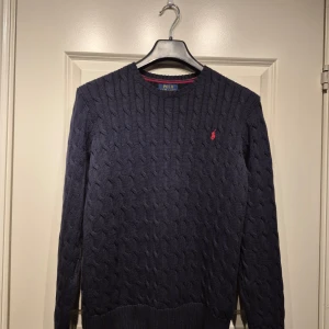 Mörkblå stickad tröja från Ralph Lauren - Mörkblå kabelsktickad tröja från Ralph Lauren.med kabelstickat mönster. Röd borderad logga på bröstet. Mycket fint skick! Hör av er med eventuella frågor eller funderingar. 