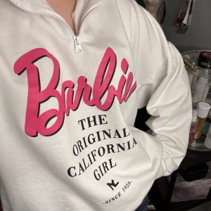 Vit Barbie-tröja med dragkedja - Säljer en vit tröja med dragkedja och tryck i rosa och svart från Barbie. Texten 'The Original California Girl' ger en retro känsla. Perfekt för en avslappnad stil.