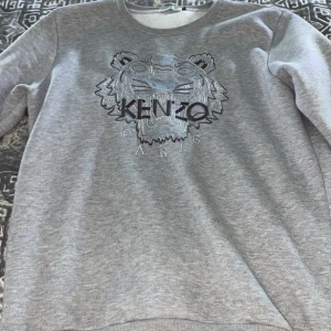 Kenzo tröja  - Säljer en stilren grå sweatshirt från Kenzo med ett broderat tigerhuvud och logotyp på framsidan. Tröjan har rund hals och långa ärmar, perfekt för en avslappnad look. Passar bra till både jeans och joggers.