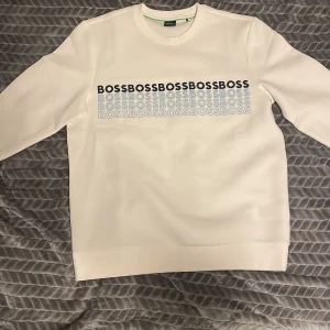 Vit tröja från Boss - Helt oanvänd Säljer en stilren vit tröja från Boss med ett tryck av märkesnamnet i svart och blått på framsidan. Tröjan har långa ärmar och en rund halsringning. Perfekt för en avslappnad och trendig look.