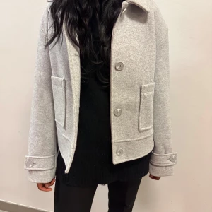 Grå kappa med knappar - Snygg grå kappa med stora knappar och bred krage. Jackan har långa ärmar och praktiska fickor framtill. Perfekt för kyligare dagar.