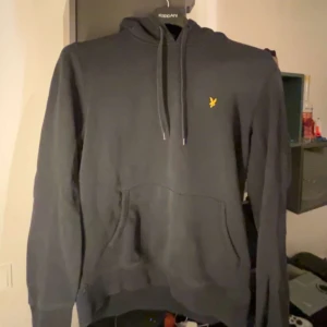 Svart hoodie från Lyle & Scott - Säljer en stilren svart hoodie från Lyle & Scott med en gul logga på bröstet. Tröjan har en praktisk magficka och justerbar huva med snören. Perfekt för en avslappnad stil.