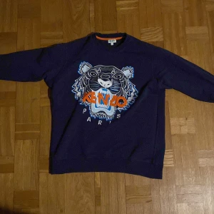 Mörkblå tröja med tigerbroderi från Kenzo - Säljer en mörkblå tröja från Kenzo med ett stort, broderat tigerhuvud i flera färger på framsidan. Tröjan har långa ärmar och en rund halsringning med orange detalj på insidan. Perfekt för en cool och avslappnad stil.