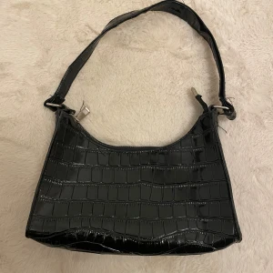 Svart axelväska med krokodilmönster - Snygg svart axelväska i syntetmaterial med krokodilmönster. Väskan har en dragkedja upptill. Perfekt för att ge en stilren touch till din outfit.