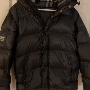 Burberry jacka  - Snygg svart dunjacka från Burberry med lång ärm och huva. Jackan har en quiltad design och stängs med knappar. Perfekt för kyliga dagar. Ganska fin men har köpt ny jacka 