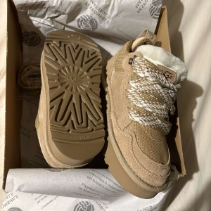 UGG - Snygga beige boots från UGG med snörning och fluffigt foder. Skorna har en robust sula och är perfekta för kyligare väder. De är tillverkade i mocka och har en stilren design med märkets logga på sidan.