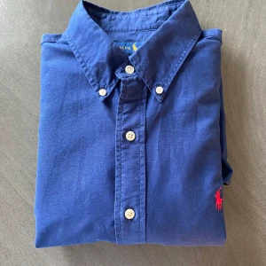 Blå skjorta från Ralph Lauren - Snygg blå skjorta från Ralph Lauren med klassisk krage och knappar framtill. Den har en broderad röd logotyp på bröstet och är tillverkad i ett bekvämt material. Perfekt för en stilren look.