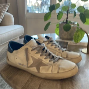 Golden goose - Snygga vita sneakers från Golden Goose med en ikonisk stjärndetalj på sidan. Skorna har grå snörning och en mörkblå häl. Perfekta för en avslappnad stil med en touch av lyx. Snabb affär = lägre pris!