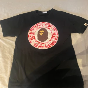 Svart t-shirt från Bape - Snygg svart t-shirt från Bape med ett stort, färgglatt tryck av deras ikoniska logga i rött och vitt på framsidan. Perfekt för streetwear-stilen. T-shirten har korta ärmar och en normal passform. Säljes i befintligt skick 