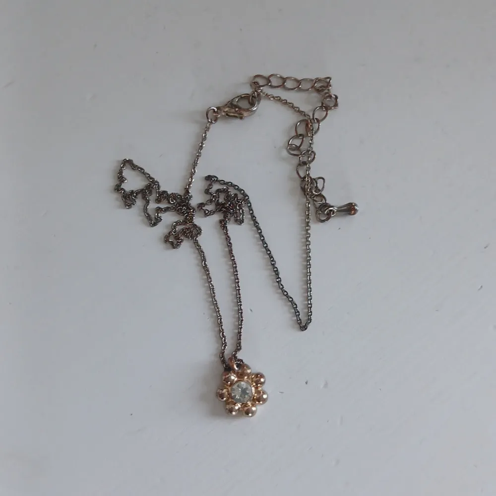Elegant halsband med en blomformad berlock i guldton. Berlocken har en glittrande sten i mitten som ger en fin detalj. Kedjan är tunn och justerbar, vilket gör det enkelt att anpassa längden. Perfekt för att lägga till en subtil touch av glamour till din outfit. Baksidan på