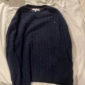Mörkblå stickad tröja från Tommy Hilfiger - Säljer en snygg mörkblå stickad tröja från Tommy Hilfiger. Tröjan har ett klassiskt flätmönster och en liten logotyp på bröstet. Står att det är 176 men passar mer som 164