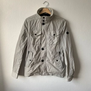 Ljusgrå Bailey jacka från J.Lindeberg - Snygg beige jacka från J.Lindeberg med hög krage och knappar. Jackan har flera fickor med knappstängning och en stilren design. Perfekt för vår och höst. Skick: Bra (dock saknas två knappar på framsidan av jackan, se bild) - Modell: Bailey Sports Nylon - Artnr: J130