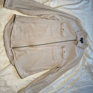 Beige overshirt från Hampton Republic - Snygg beige overshirt från Hampton Republic med dragkedja och två bröstfickor med knappar. Perfekt för lager-på-lager-stil. Den har långa ärmar och en klassisk krage.