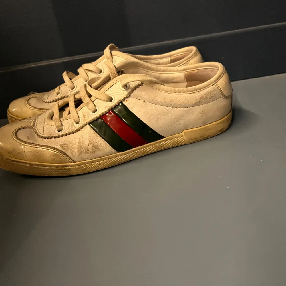Snygga beige sneakers från Gucci med klassiska ränder i grönt och rött på sidan. Skorna har snörning och är tillverkade i skinn. Perfekta för en stilren look.lite smutsiga men väldigt snygga.. Kengät.