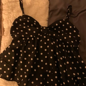 Svart prickig topp med volang - Charmig svart topp med vita prickar och volangdetaljer. Axelbandslös design med smockad rygg för en bekväm passform. Perfekt för en somrig look.