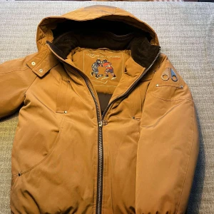 Brun jacka från Moose Knuckles - Säljer en snygg brun/beige jacka från Moose Knuckles med huva och dragkedja. Jackan har coola metalliska detaljer på ärmen och en quiltad insida. Perfekt för kyliga dagar med sin varma vaddering. Den används inte längre. Storlek XL i BARNSTORLEK. Skulle säga XS eller XXS i man storlek. Nyskick, absolut inga täcken på användning. Vid frågor hör av er, mvh Maxi