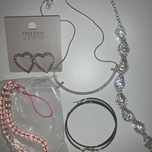 Smyckeset - Ett set med glittriga smycken från Freedom by Topshop. Innehåller hjärtformade örhängen, två halsband med strassdetaljer och två armband, ett i rosa och ett i silver. Perfekt för att lägga till lite extra bling till din outfit.