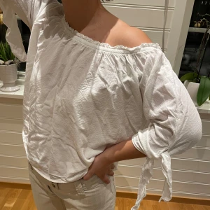 Vit offshoulder blus - Säljer en stilren vit offshoulder blus med knytband vid ärmarna. Perfekt för en avslappnad och chic look. Passar bra till både jeans och kjol.