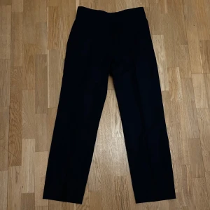 Mörkblå kostymbyxor från Riley - Snygga mörkblå kostymbyxor från Riley. Byxorna har en klassisk design med knappar och bälteshällor, perfekta för en stilren look. Avslappnad passform. Endast använda två gånger. 