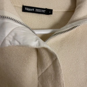 Beige stickad jacka från Filippa K - Säljer en helt ny stilren beige stickad jacka från Filippa K med dragkedja och quiltade detaljer. Jackan har långa ärmar och en bekväm passform, perfekt för kyligare dagar. Den är designad i Sverige och producerad i Kina. Går att använda som en tunn jacka under sommar och våren, 