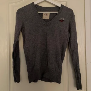 Grå V-ringad tröja från Hollister - Säljer en stilren grå V-ringad tröja från Hollister. Tröjan har långa ärmar och en liten broderad logga på bröstet. 250kr🤌🏼 Endast testad, ej använd.