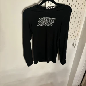 Svart långärmad tröja från Nike - Säljer en svart långärmad tröja från Nike Pro med vit logga på bröstet och text på baksidan av kragen. Perfekt för träning eller vardagsbruk. Materialet är mjukt och stretchigt för optimal komfort.