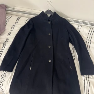 Svart kappa från Schneiders - Elegant svart kappa från Schneiders i storlek 38. Klassisk design med knappar framtill och diskreta fickor med dragkedja. Perfekt för kyligare dagar med sin stilrena look och långa ärmar. En tidlös investering för garderoben.