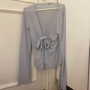 Ljusgrå långärmad topp med knyt - Säljer en stilren ljusblå topp med långa ärmar och en snygg knytning framtill. Toppen har en djup v-ringning och är tillverkad i ett mjukt material som ger en skön passform. Perfekt för en chic och avslappnad look. Köptes från Gina för 300 och är knappt använd💞